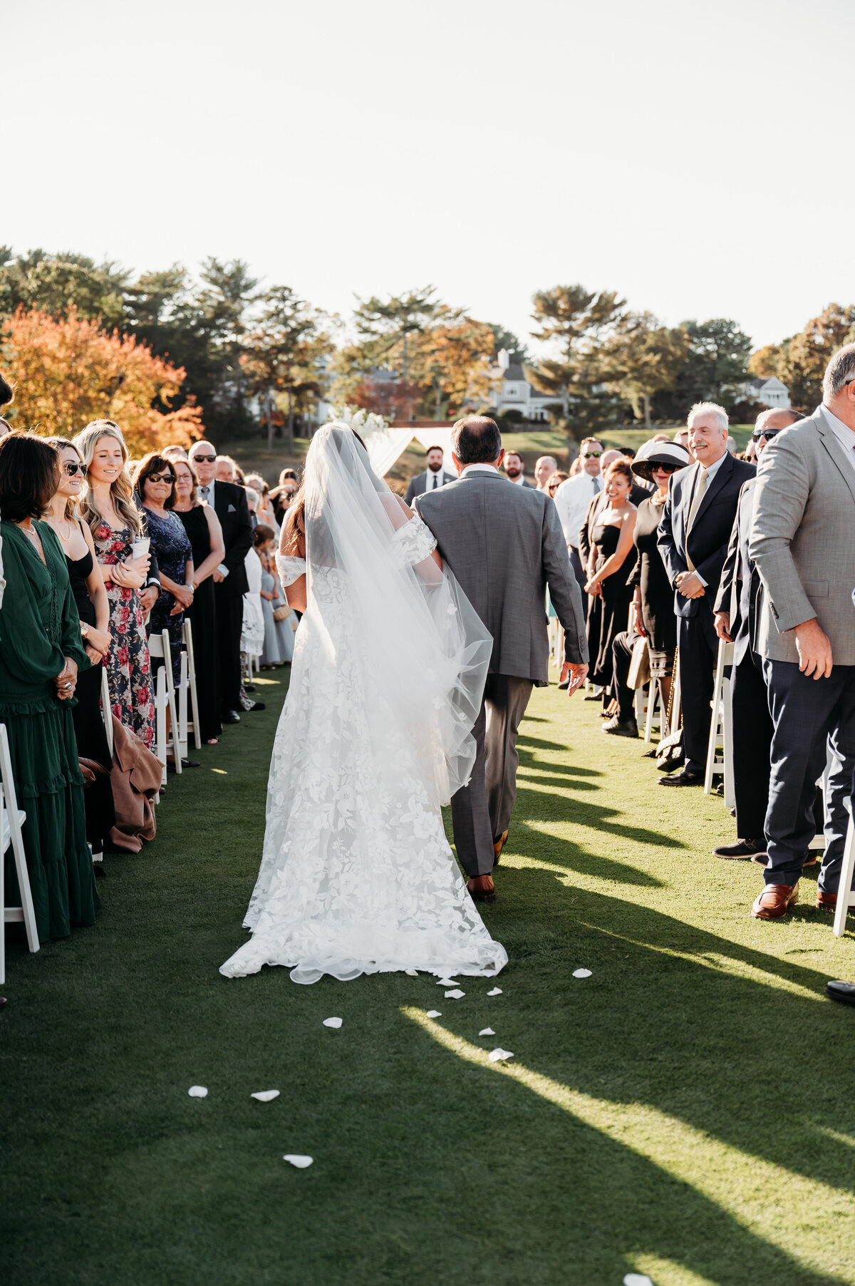 Emotional outdoor wedding ceremony as a bride walks down the aisle with her father, surrounded by smiling guests and golden afternoon light. Her flowing veil and elegant lace gown create a timeless, romantic moment. Captured on a scenic fall wedding day, this image beautifully showcases the joy and anticipation of saying 'I do.' Captured by Moll – Boston wedding photographer specializing in heartfelt and documentary-style wedding photography.