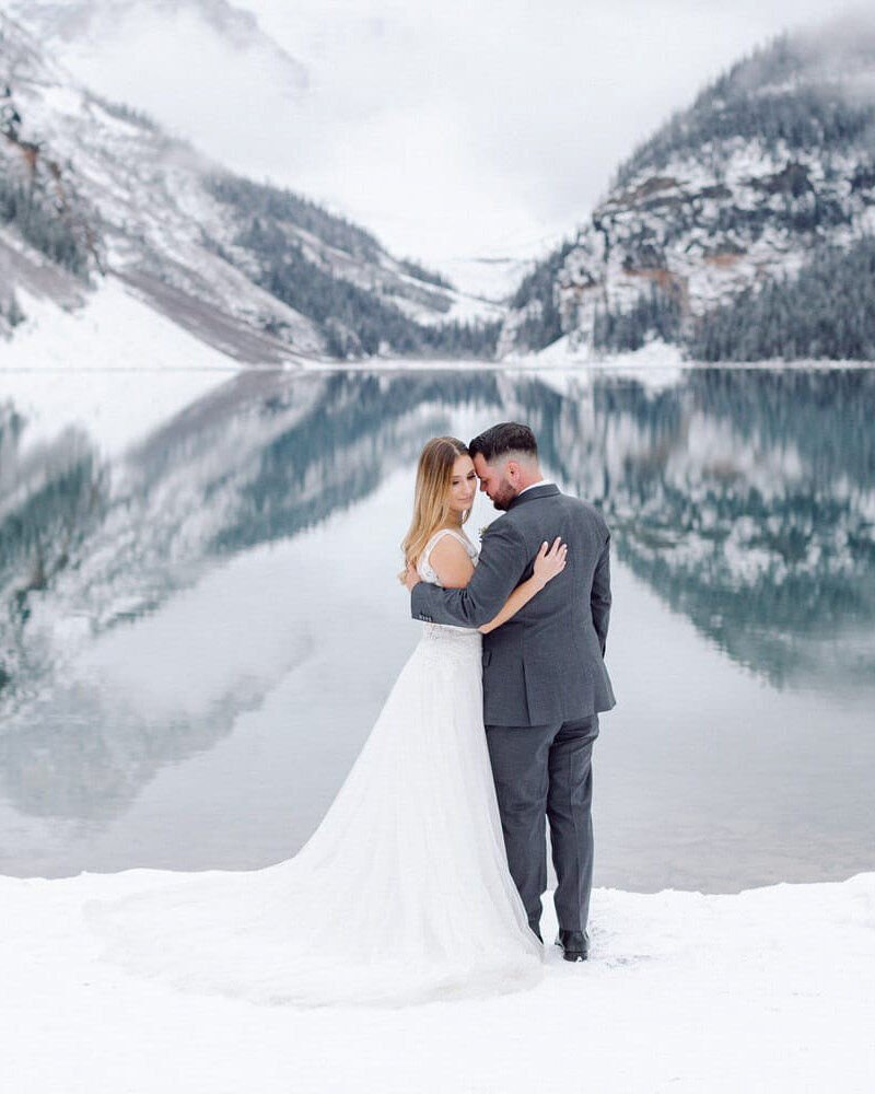 This breathtaking photograph captures the bride and groom holding each other on the shoreline of Lake Louise during the fall, surrounded by fresh snow and a perfectly still, reflective lake. After an evening stroll from their wedding reception at the Fairmont Chateau Lake Louise Hotel, the scene radiates serenity and romance.