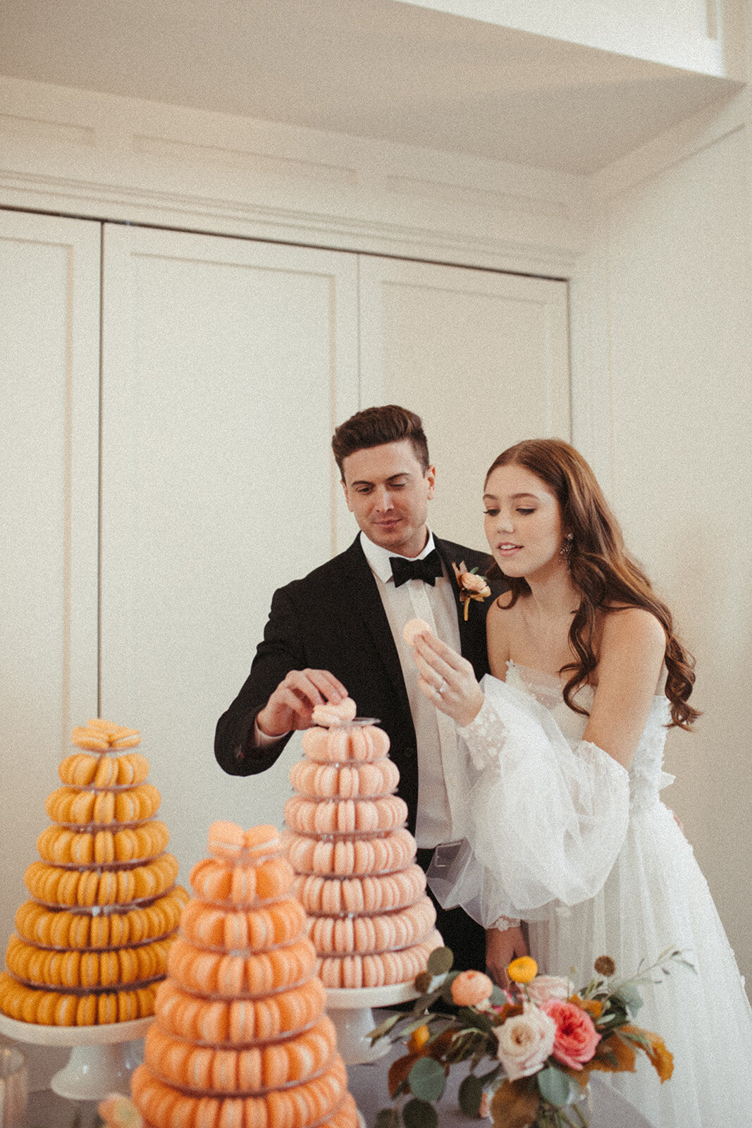 A bride and groom wearing a white wedding gown and black tuxedo lean into a dessert table filled with macarons.