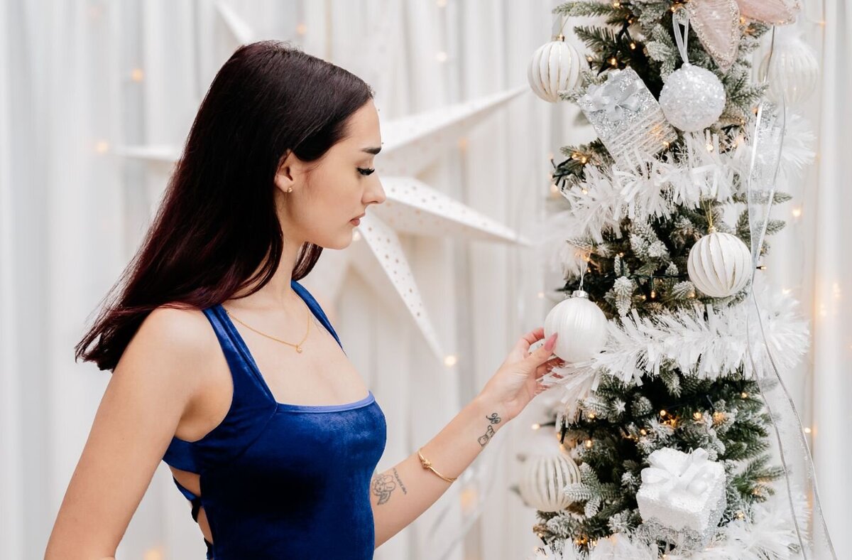Woman decorating Christmas tree – Young woman in a blue velvet dress hanging a white ornament on a sparkling white Christmas tree.