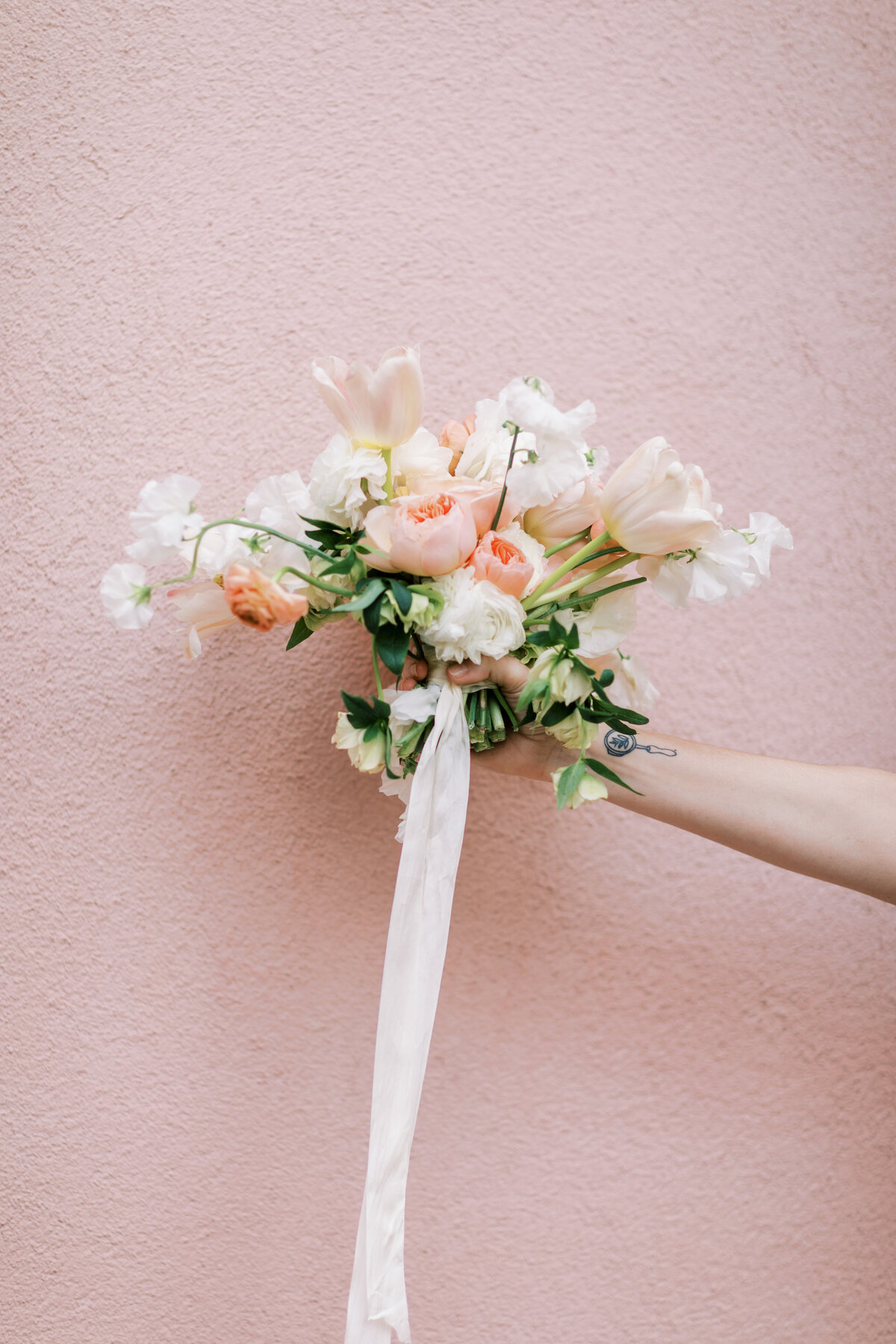 White and pink wedding bouquet with a long pale pink ribbon dangling being held by an arm in front of a pastel pink wall on Rainbow Row in Charleston