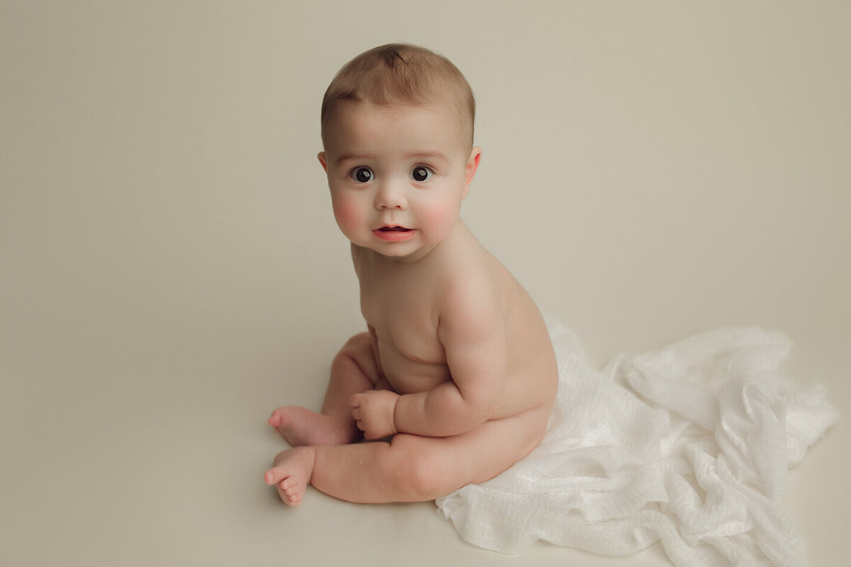 a six month old baby sitting nude on a white fabric on a white backdrop
