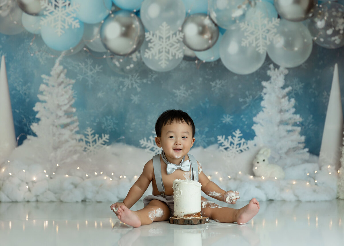 A happy toddler boy sits in a white christmas themed New Jersey Cake Smash Photographer studio for his first birthday