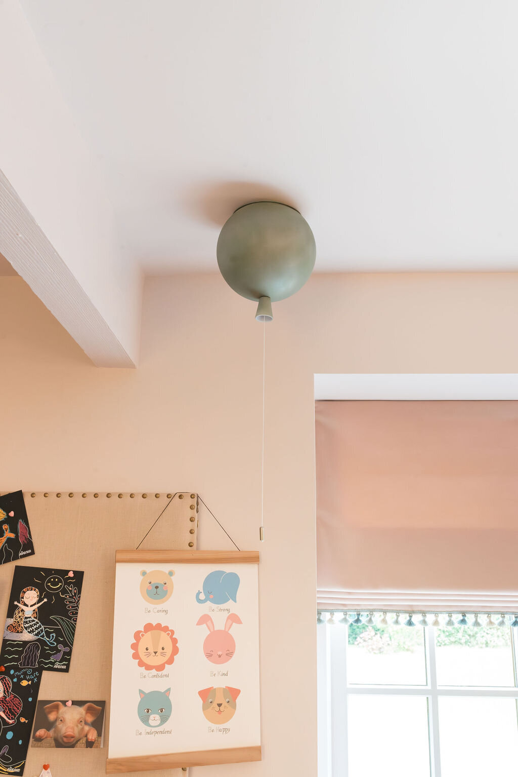 A children's pastel playroom with a window and a board showcasing children's artwork. A green balloon art hangs from the celing.