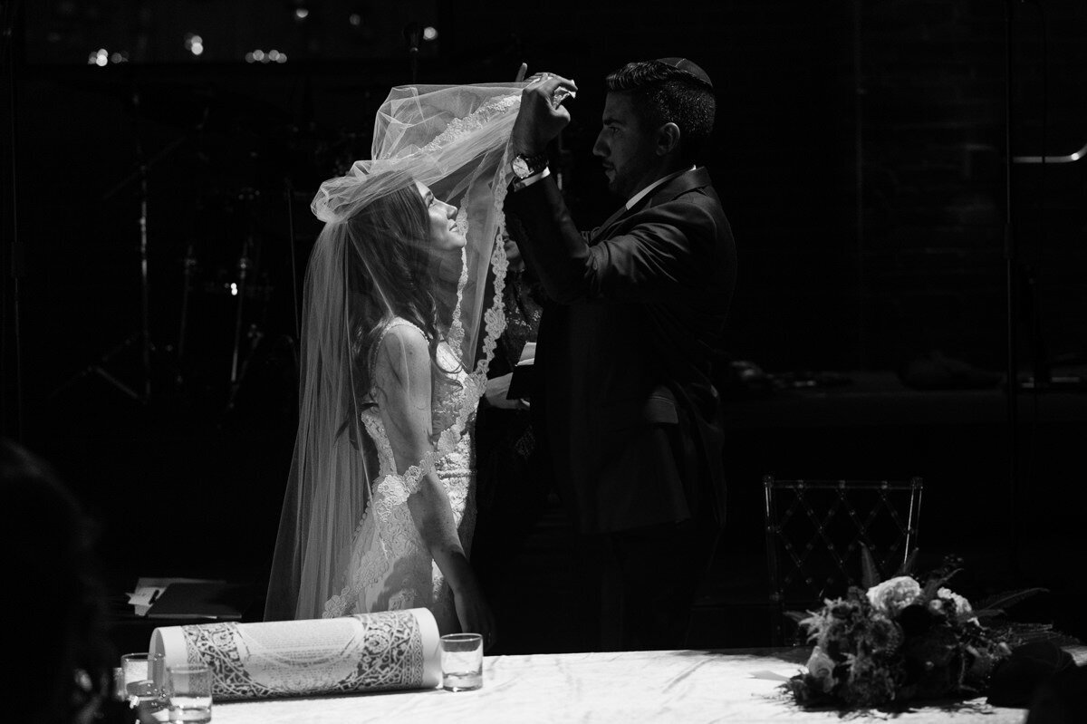 Black and white image of a bride on her wedding day looking up to the light that illuminates her face while her to-be husband veils her at the bedecken