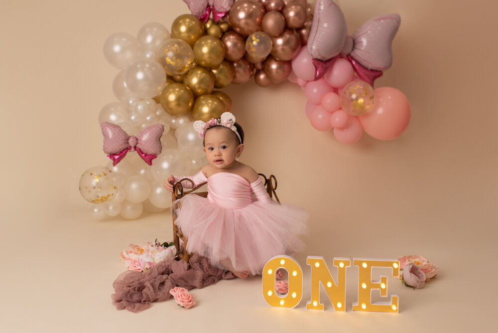 Atlanta, Georgia cake smash photography session baby girl wearing pink dress with tute and minnie mouse ears sitting on bench with gold and pink balloon garland in the backdroung