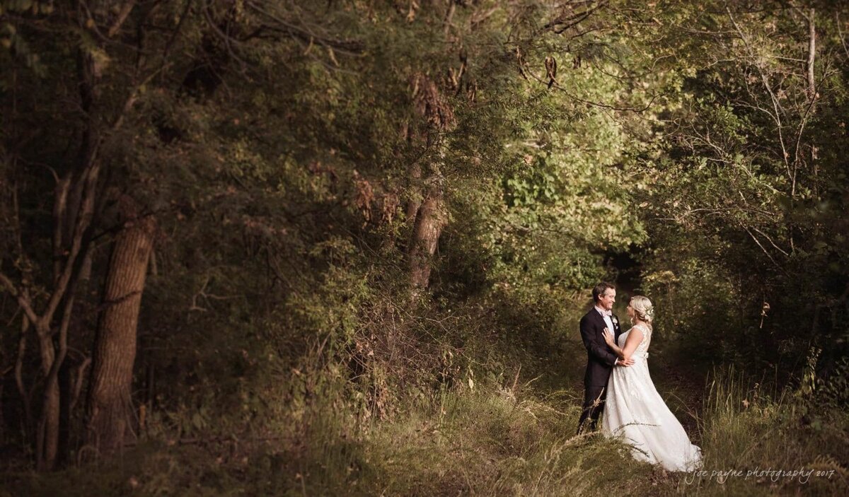 A bride and groom standing close and smiling in the woods.