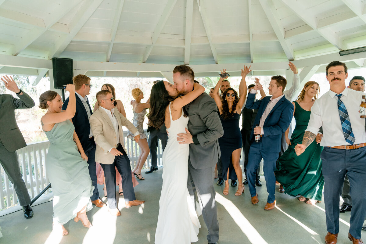 bride and groom in the middle of the dance floor kissing while guests are surrounding them dancing