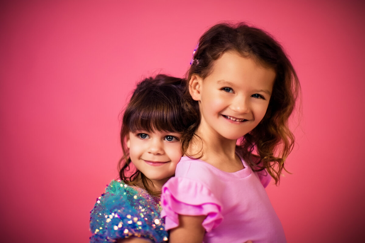 Sisters embrace and look off into the distance with their bright blue eyes their colorful outfits at a styled Studio session at the Pittsburgh Studio on Pink backdrop