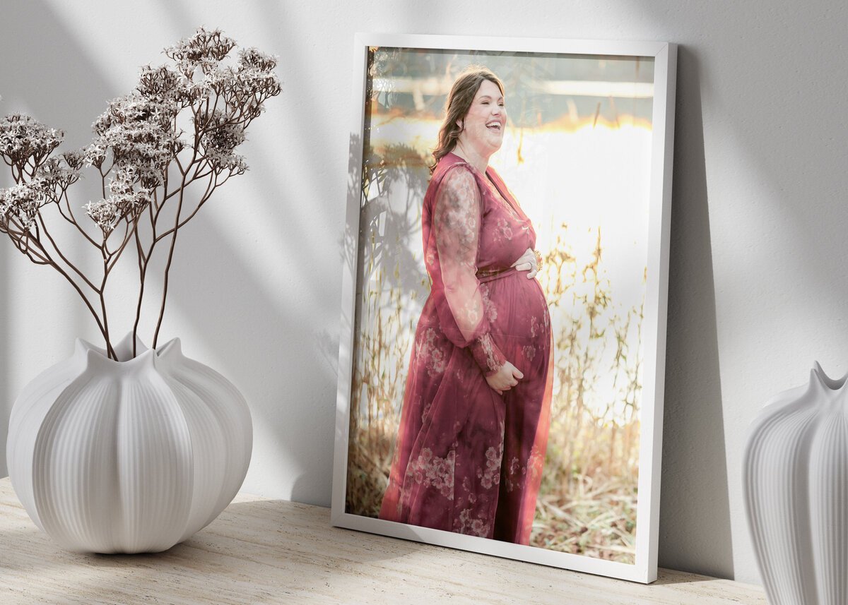 A pregnant woman, who is wearing a red floral dress, laughs during her maternity session.