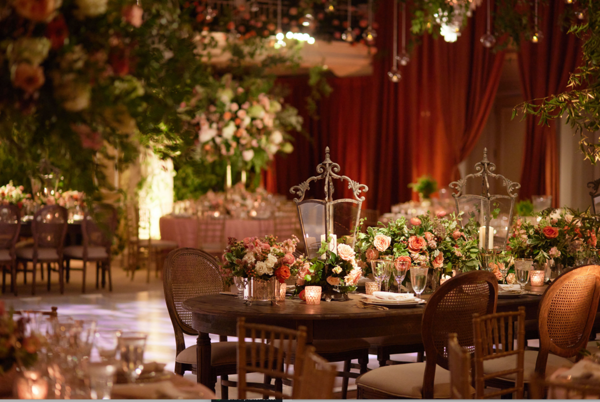 This is where the photo was taken: Washington D.C., The Willard Intercontinental, one of the top wedding venues and hotels in DC by photographer Shawn Connell, with Clayton Films as videographers. The venue features a luxuriously decorated indoor event space with elegantly set tables adorned with floral arrangements, candles, and fine tableware. The ambiance is warm, with soft lighting, rich red curtains, and greenery hanging from above, creating a sophisticated atmosphere.