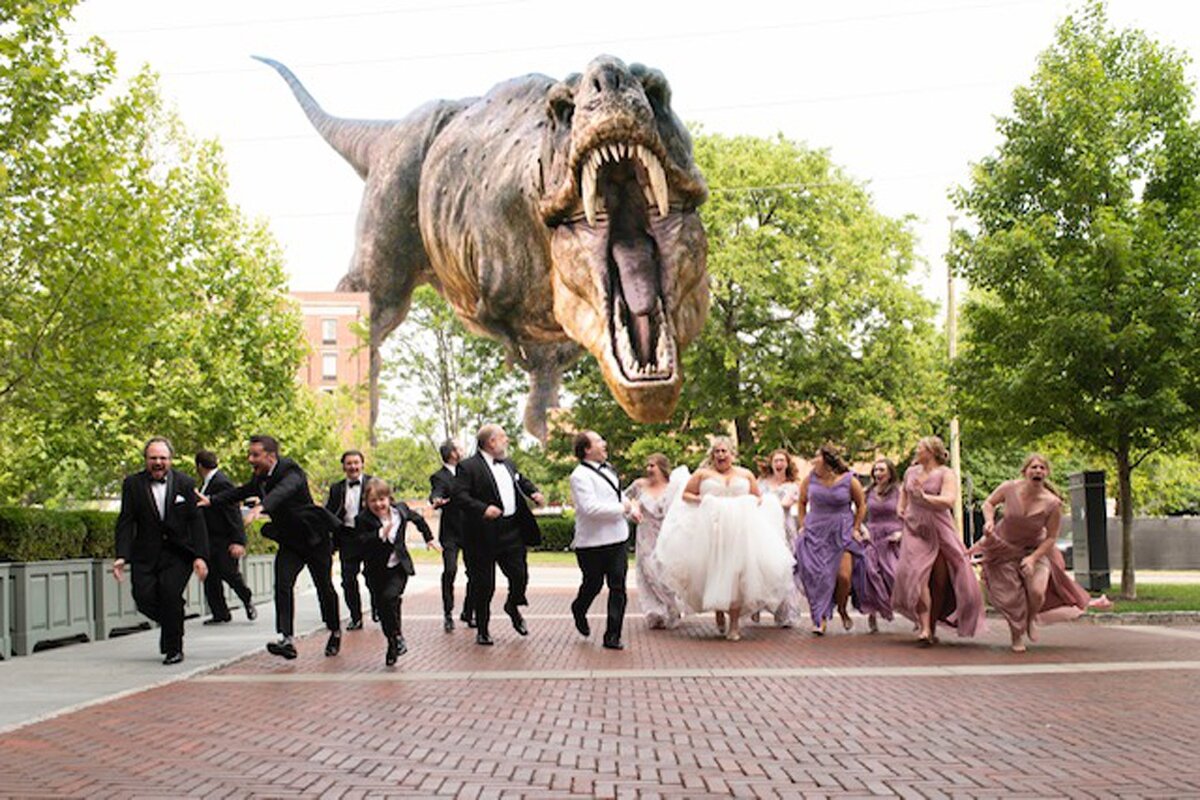 The groomsmen wearing black tuxedos and the bridesmaids wearing purple dresses run alongside the bride in a flowing white dress and the groom in a tuxedo with a white jacket as they are bring chased by a large t-rex in Nashville.