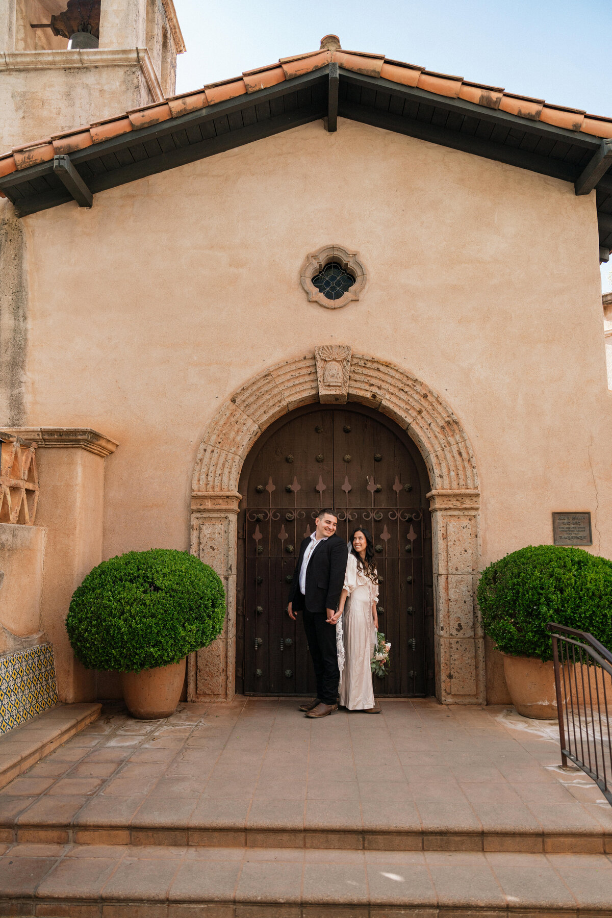 Bride and groom holding hands in front of the iconic chapel doors at Tlaquepaque Arts & Shopping Village in Sedona, Arizona, surrounded by desert-inspired architecture and lush greenery during their intimate elopement.