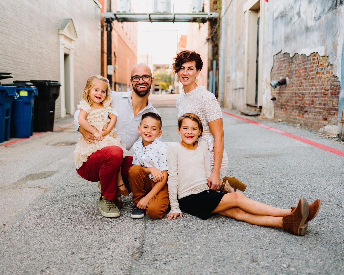 Professional mini photos session for a actual family in plano,texas. The father is holding his baby girl and is wearing a red pant and white t-shirt. The children around him are sitting on the street floor and are looking at the camera