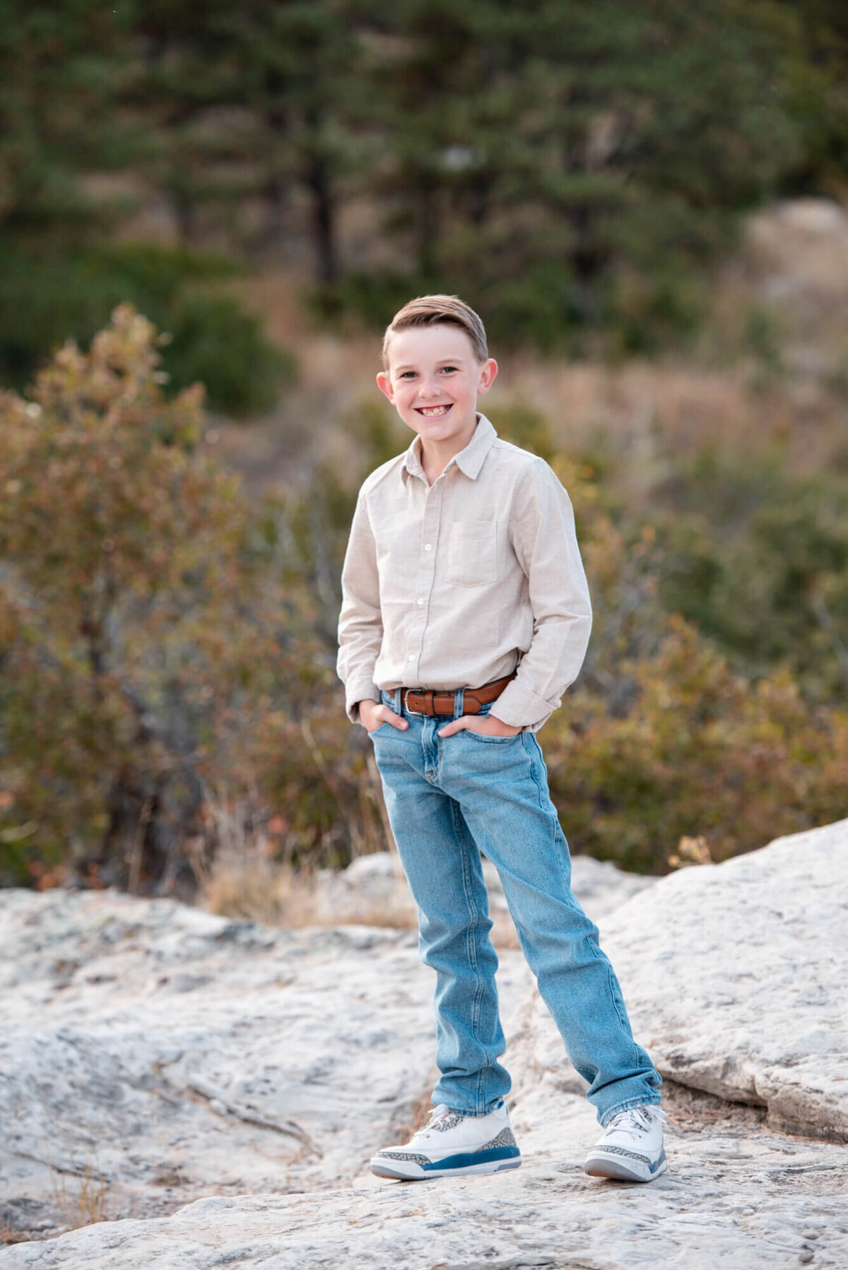 A young boy stands with hands in jean pockets while smiling on a large boulder