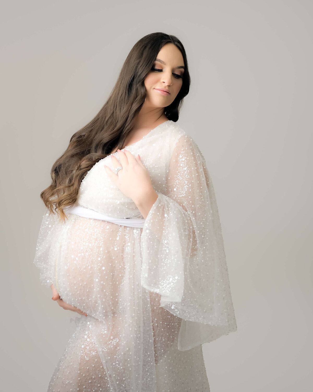 photo by E. Benecki Photography of an expecting mother in a white sequin sheer robe, set against a white background, her long brown hair flows beautifully as she looks over her shoulder.