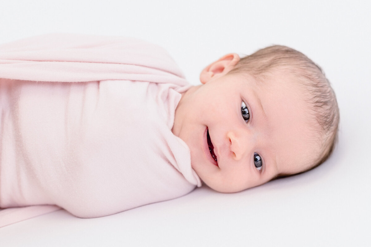 A baby wrapped in a soft, light pink blanket lies on a white surface. The baby has light hair, with blue eyes, and is smiling gently while looking slightly to the left.