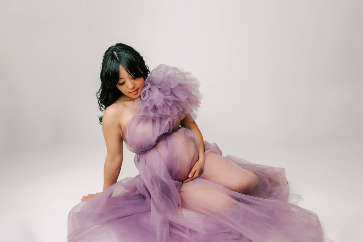 woman with black hair laying on white backdrop with her hand on her belly. She is wearing a purple couture gown with a poof on one shoulder.
