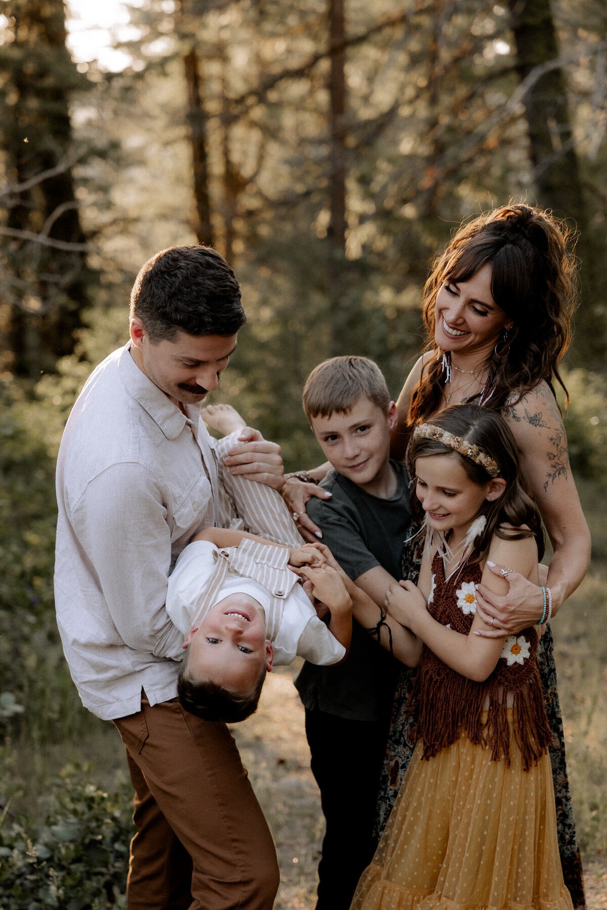 Golden sunlight glow shining through the trees. Mom is in a boho dress with her arms wrapped around her son and daughter. Dad is joyfully playing with the youngest son. Capture by Mary Wiebe photography