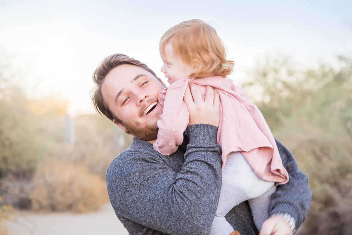 brown haired man in gray shirt holding and laughing with redheaded toddler niece in pink dress at desert park