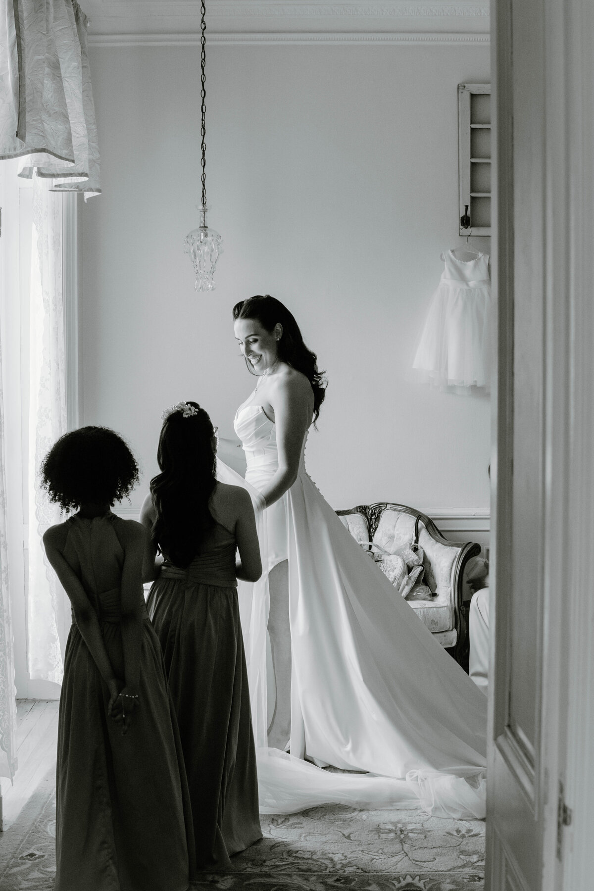 Newport Wedding Photographer | A bride in an elegant gown smiles warmly at two bridesmaids in a softly lit room, conveying joy and anticipation. A delicate dress hangs on the wall.