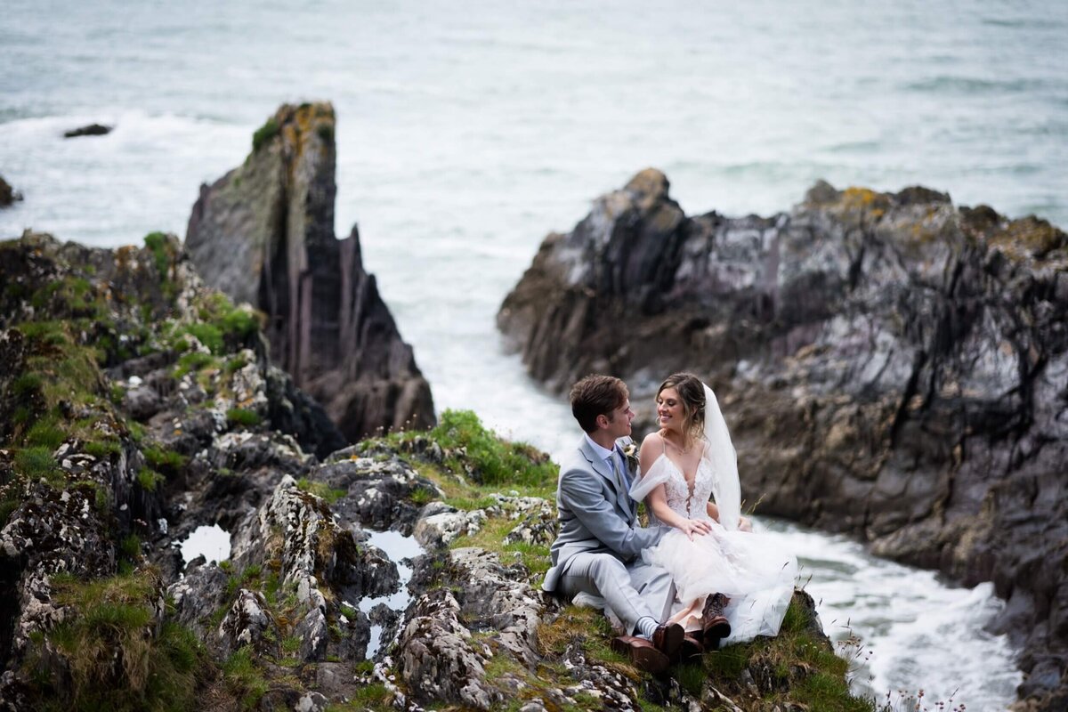 A bride and groom sitting on a rock by the sea