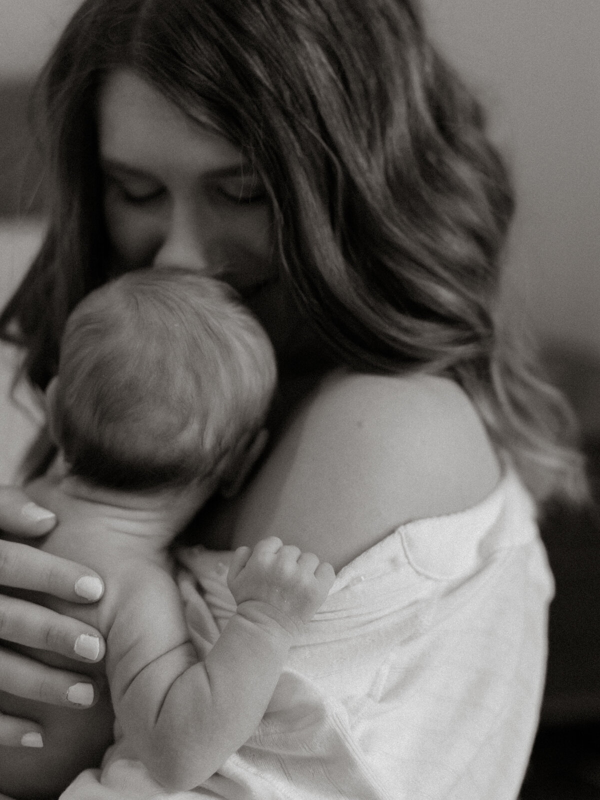 Black and white photo of a mother holding her diapered newborn facing her, gently smelling or kissing his face during a quiet in-home session filled with connection.