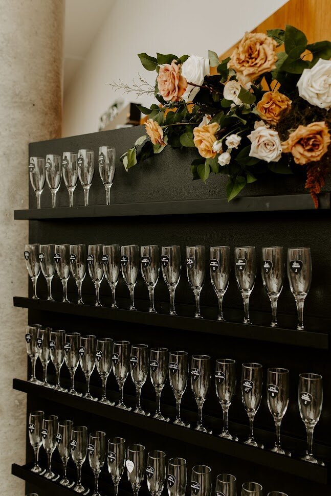 This close-up showcases a stunning black champagne wall adorned with lush earth-tone florals at the top. Each filled champagne flute is elegantly paired with unique black guitar pick-shaped escort cards, perfectly reflecting the groom’s rock band theme. This chic detail is ideal for couples looking to add a personal touch to their wedding decor in Ottawa.