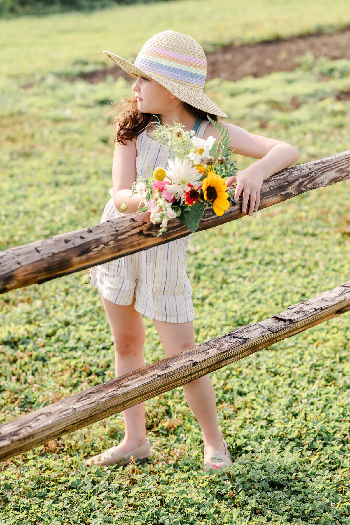 A young girl, wearing an  off white romper and sun hat leans on a fence while holding a bouquet of flowers.