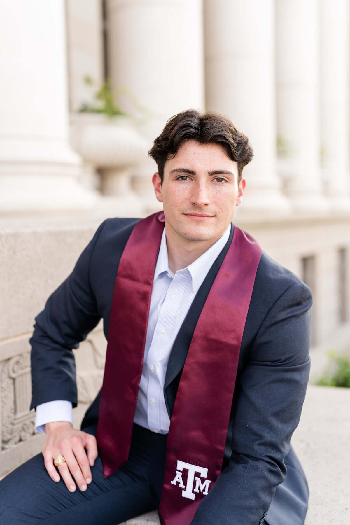 Texas A&M senior guy sitting in front of Administration Building and leaning on leg while wearing suit and Aggie stole