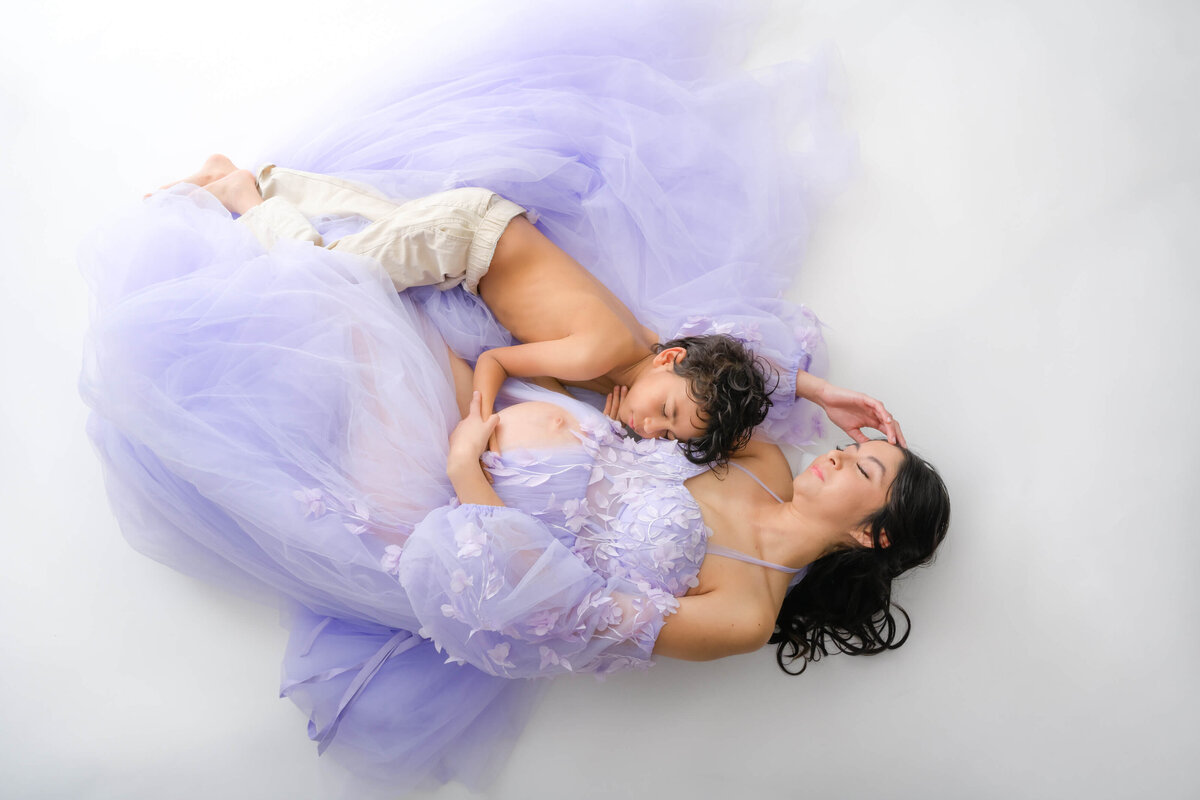 Beth Benecki of E. Benecki Photography photographs a maternity photo session with a white background.  Expecting mother is wearing an off the shoulder chiffon lavender dress.  She is laying on the floor with her son snuggled close to her  and hugging her glowing belly.