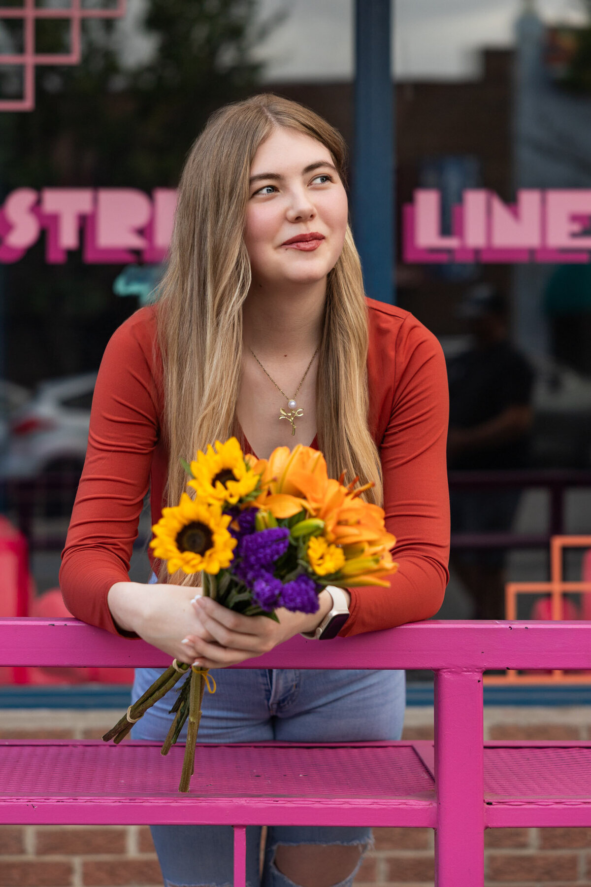 A senior girl hold sunflowers leaning against a pink railing in downtown Lawrence, KS