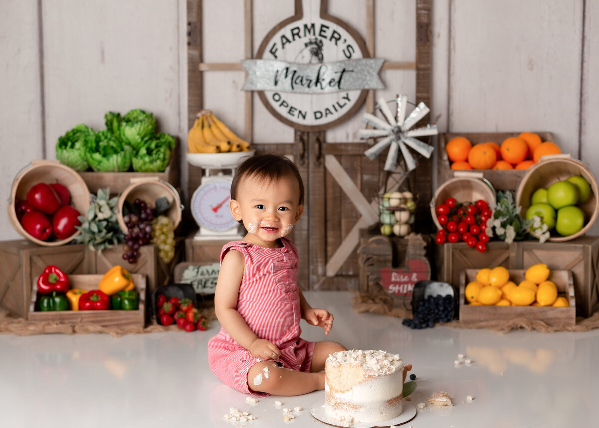 Farmers Market themed cake smash in West Palm Beach and Jupiter newborn and cake smash photography studio.  Baby girl in pink outfit sitting in front of white cake with cake on her face and smiling at the camera. In the background there is as vintage barn door and produce baskets of colorful fruit and vegetables.