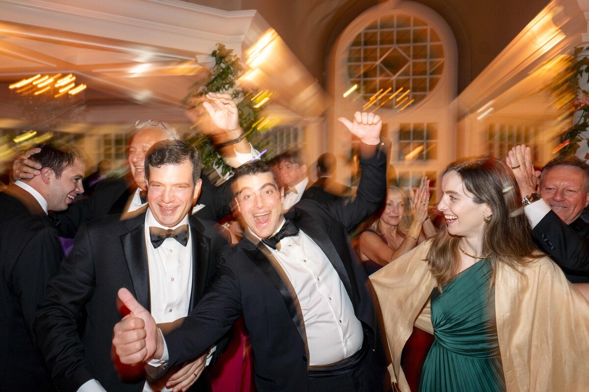A group of people in formal attire are celebrating indoors. Two men in suits are joyfully raising their arms, while a woman in a green dress with a shawl smiles. The background features warm lighting and large windows.