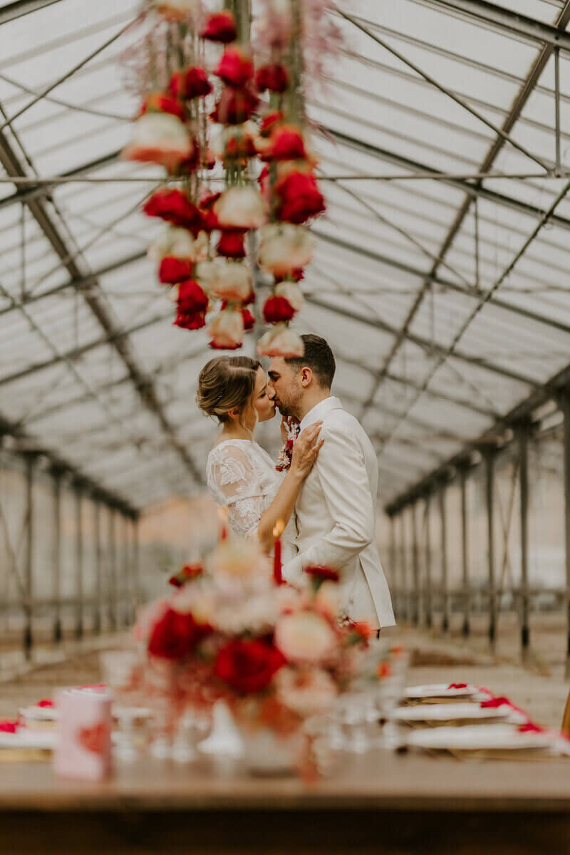 Mariés s'embrassant avec au premier plan une table en bois décorée pour un mariage et des fleurs suspendus rouges et blanches à une verrière. Séance photo réalisée pour le workshop photographie de mariage.