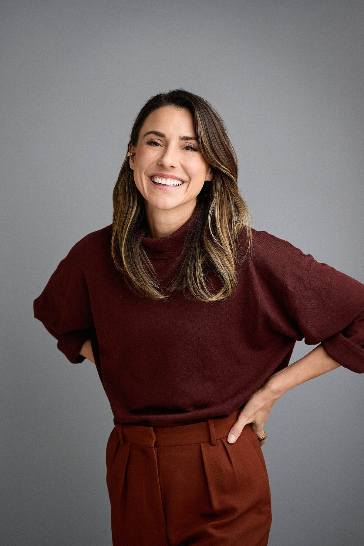 Smiling woman with hands on hips wearing burgundy jumper and rust trousers. Against a grey studio background.