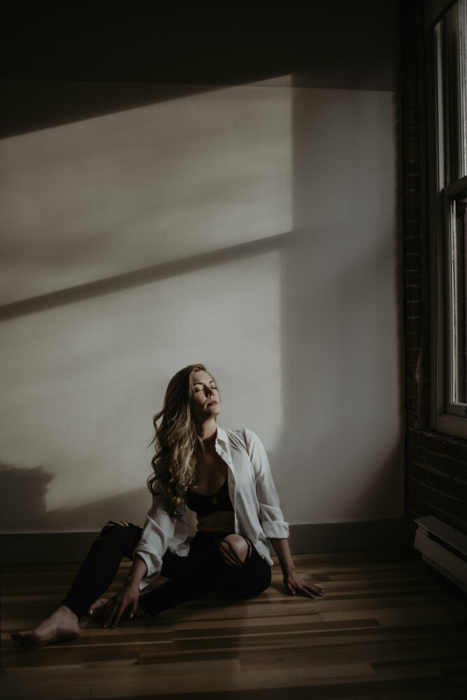 girl sitting on the floor in an empty room. There is a window letting in direct light that hits the wall behind her in large rectangles. She is sitting with her legs out and tilting her head back to face the window. Eyes are closed and she is embracing the light.