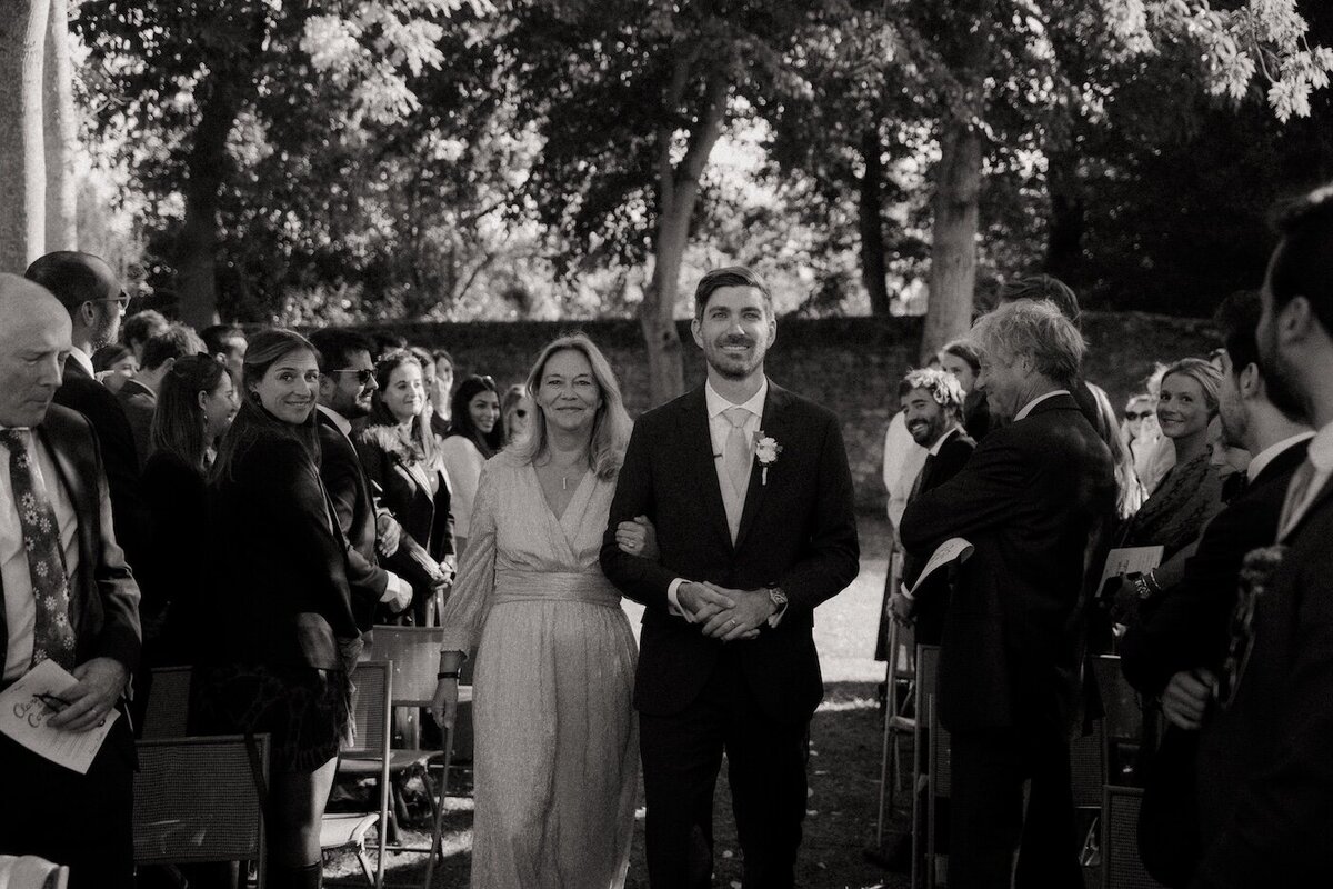 Le marié remonte l'allée du jardin au bras de sa mère, entouré des invités lors de la cérémonie. Photographie de mariage en noir et blanc signée Kamescop.