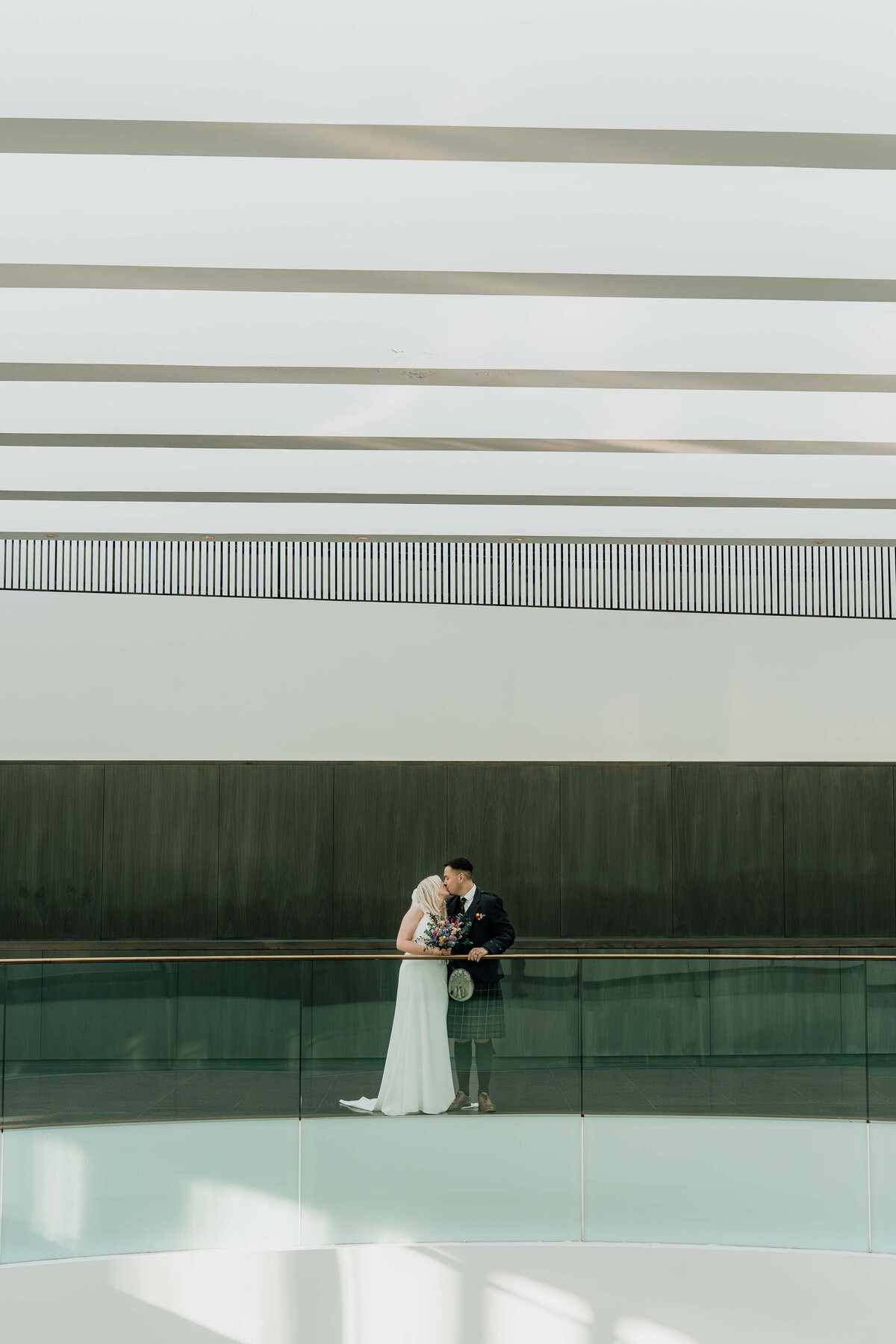 A couple kiss in a large modern space in Aberdeen Art Gallery. They are photographed from a distance by their Aberdeen wedding photographer.