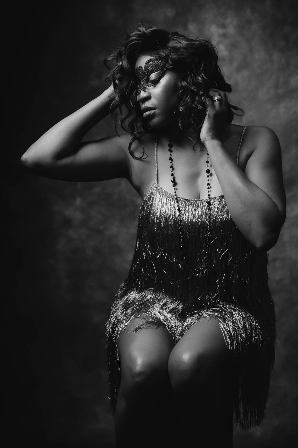 a black and white portrait of a woman wearing an eye mask sitting on a stool touching her hair