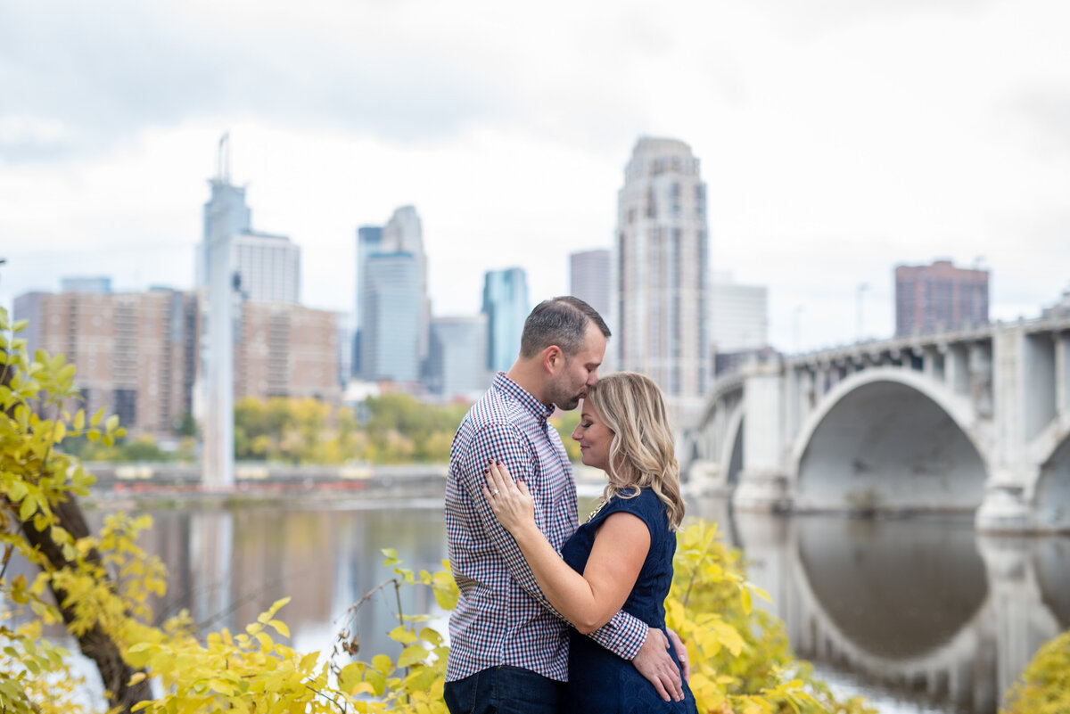 Engaged couple embrace as man kisses his future bride's forehead in front of city skyline