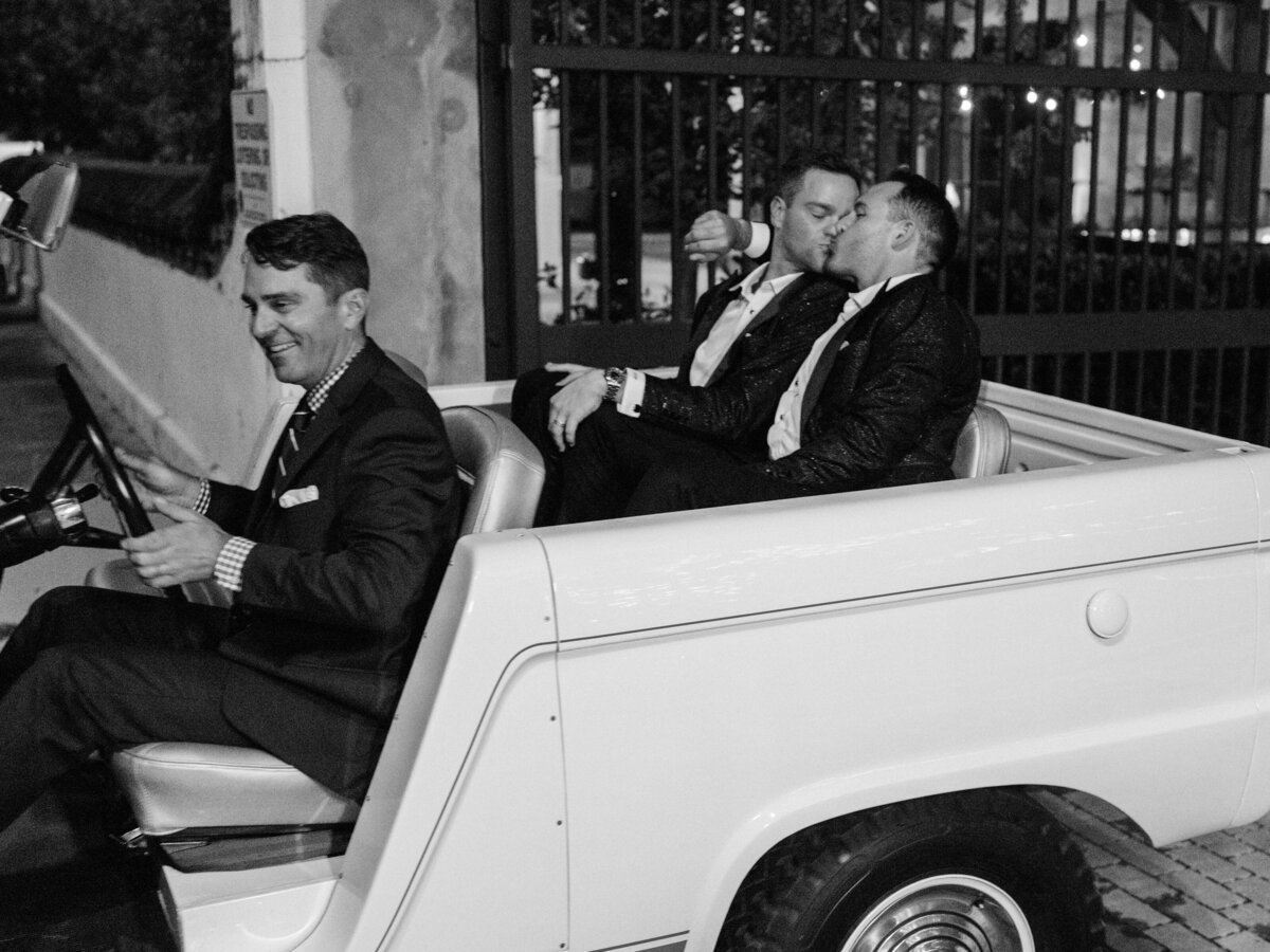 A tender kiss shared by a couple in a classic vehicle as they depart, encapsulating the joy and love of their special night and memories with friends.