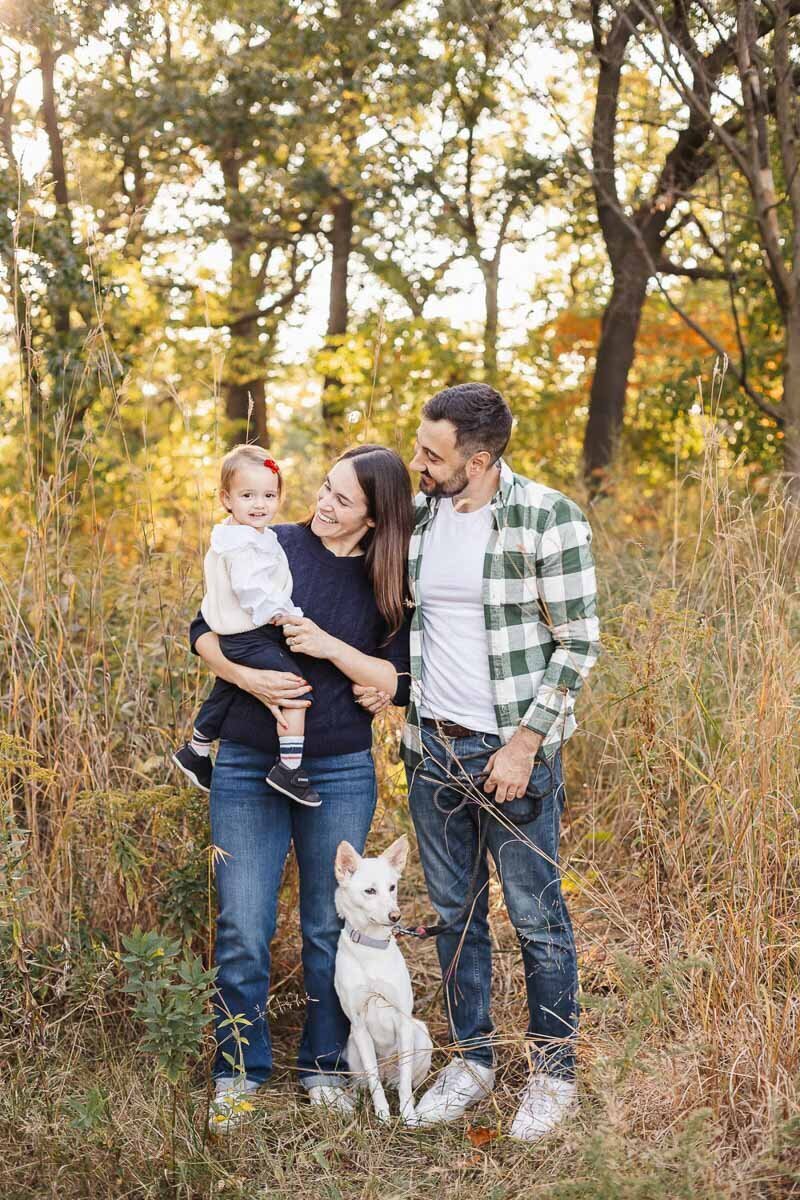 A family of three and their white dog enjoy a sunny day outdoors in a grassy area surrounded by tall trees. The woman cradles a child while the man sports a green plaid shirt, capturing the essence of natural Toronto family photography.