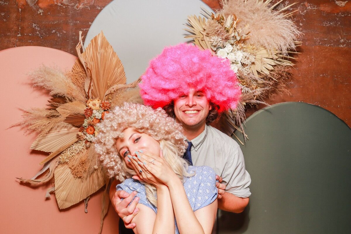 couple posing with wigs during a wedding reception entertainment, with modern arches in the background, perfect for photo booth setups