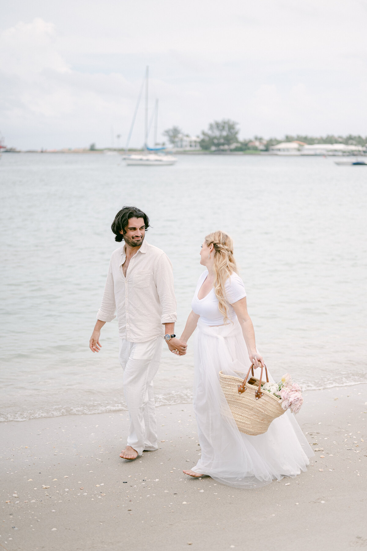 Bride holding a French basket with flowers, holding her husband's hand on the beach