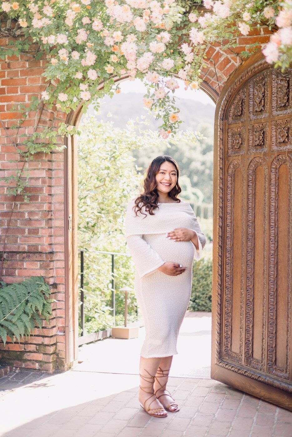 Pregnant woman cradling belly while gazing at camera, framed by a beautiful arched doorway with lovely flowers during Filoli Gardens maternity session