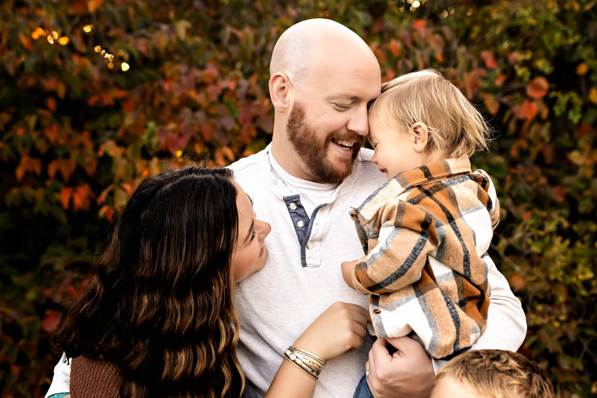A smiling man holds a young child in a plaid shirt as they pose for outdoor portraits. A woman with long dark hair embraces them, looking up fondly amidst the autumn foliage, capturing a heartfelt moment from Bloomington family photographer.