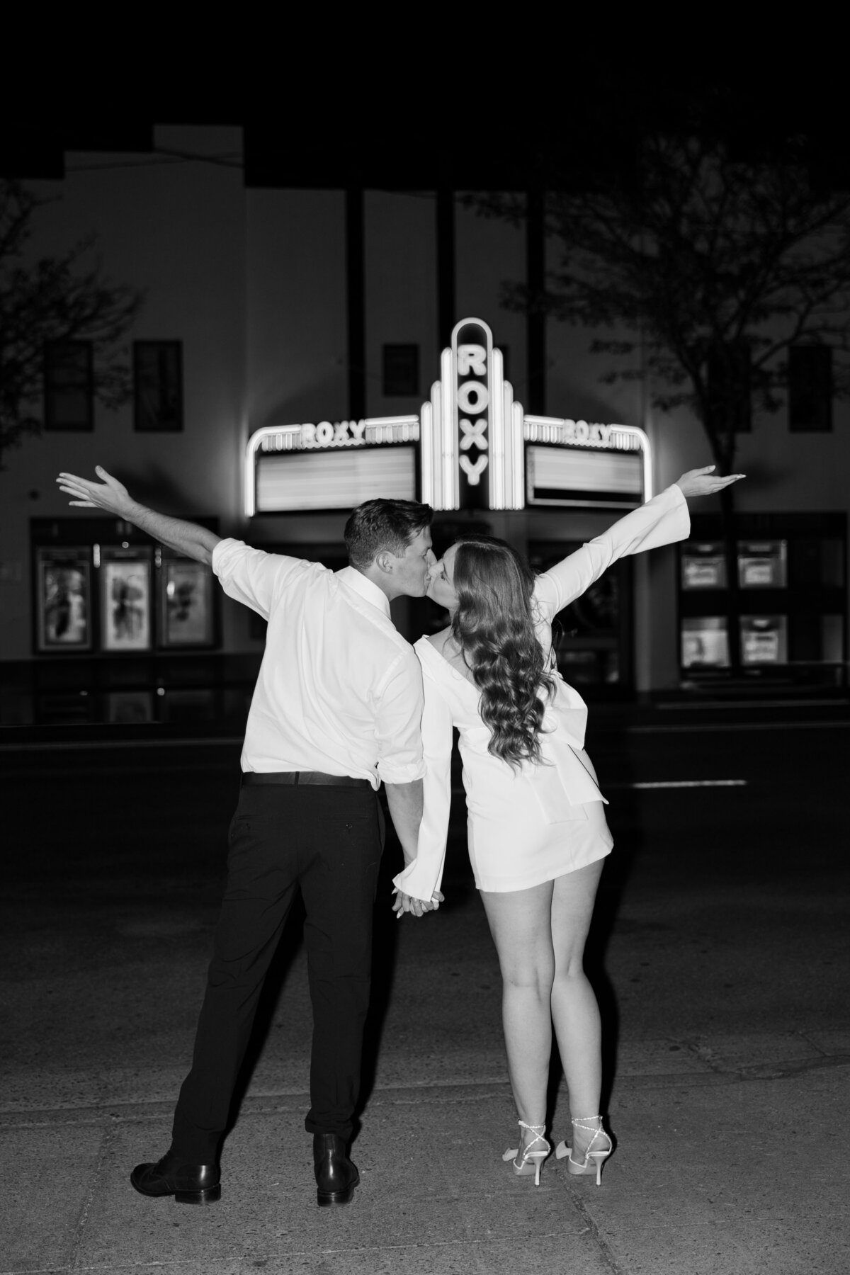 Black and white photo of a couple kissing in front of a vintage movie theater.