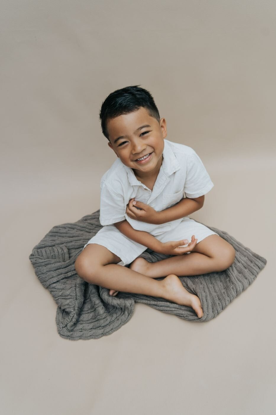  A fine art portrait of a young boy sitting cross-legged on a gray knit blanket. He is wearing a light-colored polo shirt and shorts and is looking up at the camera with a wide, joyful smile. The background is a simple, solid tan color.