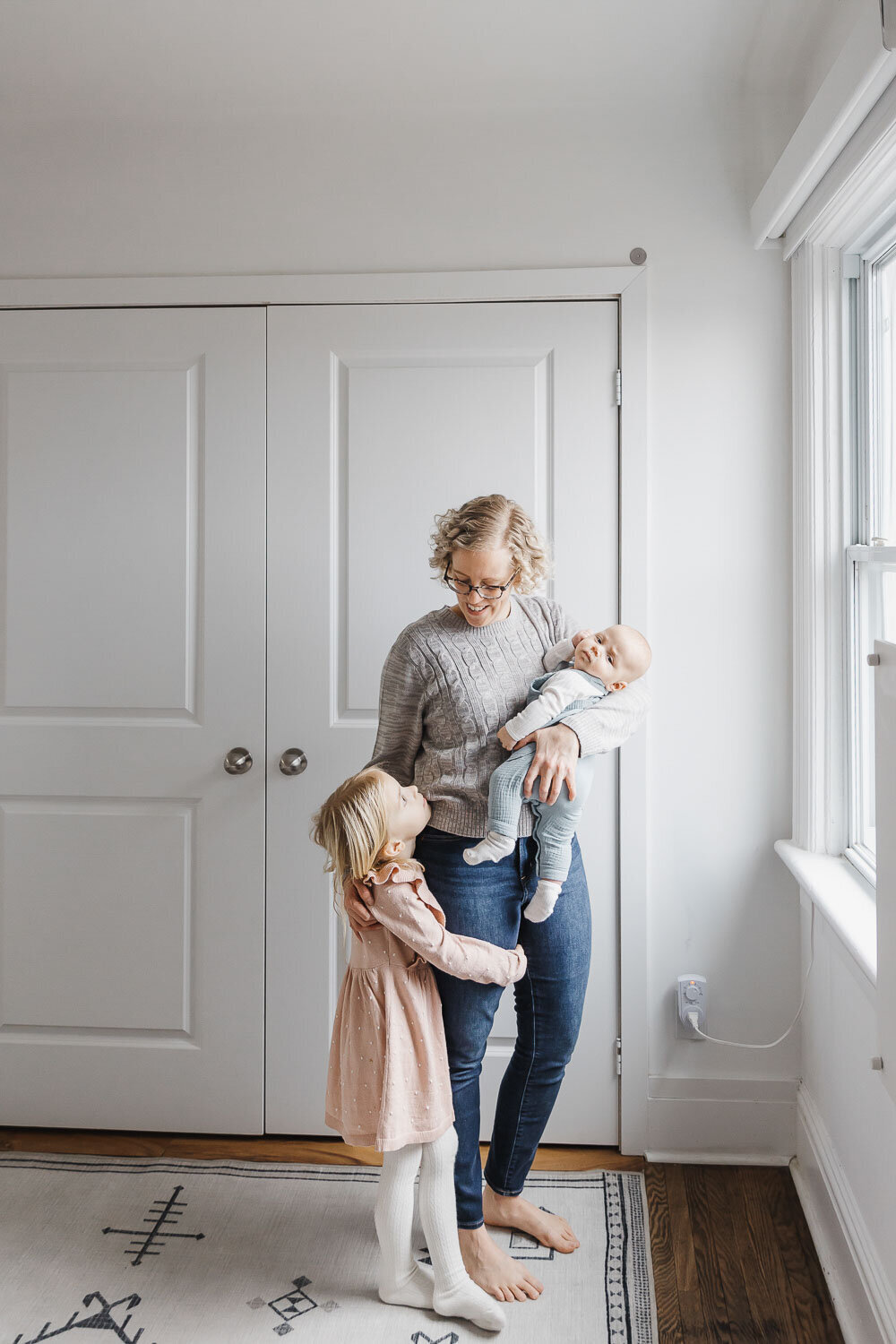 Mom holding newborn son while big sister hugs moms leg.